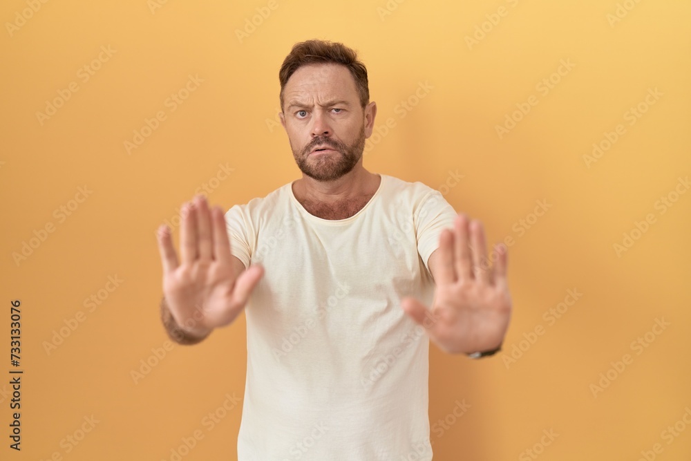 Middle age man with beard standing over yellow background moving away hands palms showing refusal and denial with afraid and disgusting expression. stop and forbidden. Middle age man with beard standing over yellow background moving away hands palms showing refusal and denial with afraid and disgusting expression. stop and forbidden.