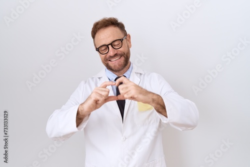 Middle age doctor man with beard wearing white coat smiling in love doing heart symbol shape with hands. romantic concept.