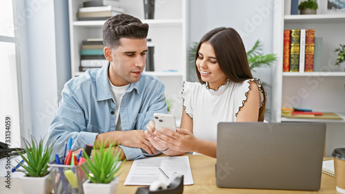 Man and woman, coworkers, sharing a smartphone in a modern office environment, with a laptop and documents on the desk.
