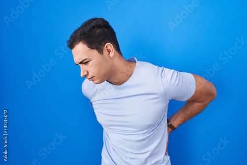 Young hispanic man standing over blue background suffering of backache, touching back with hand, muscular pain