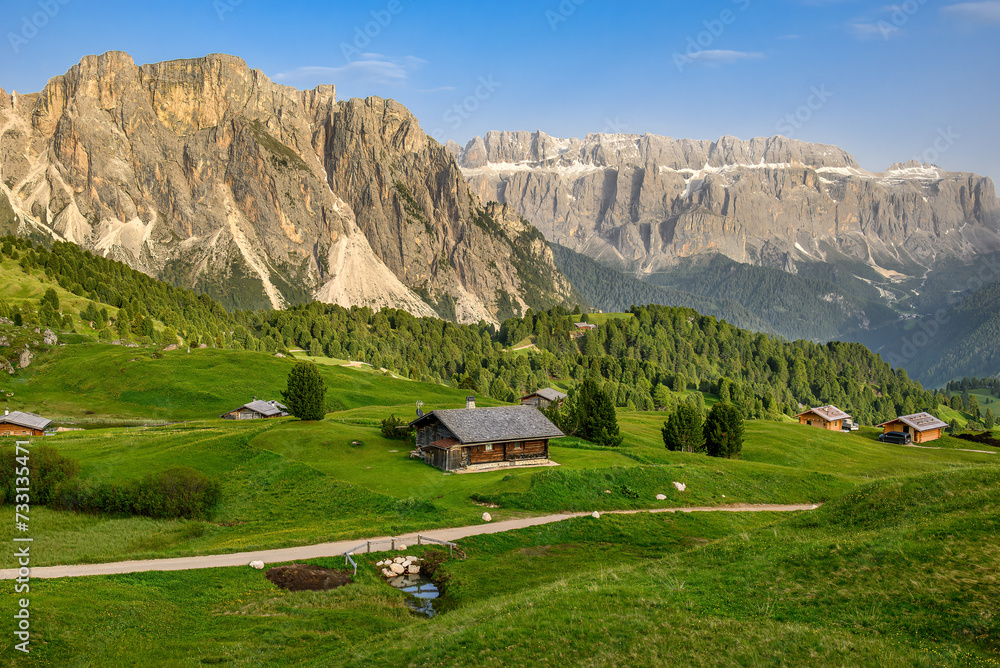Alpine huts on the grassy slopes of the Italian Dolomites with rocky ranges in the background.