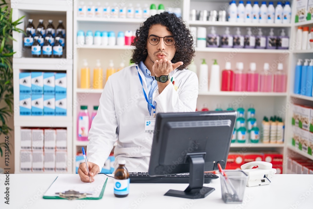 Hispanic man with curly hair working at pharmacy drugstore looking at the camera blowing a kiss with hand on air being lovely and sexy. love expression. Hispanic man with curly hair working at pharmacy drugstore looking at the camera blowing a kiss with hand on air being lovely and sexy. love expression.