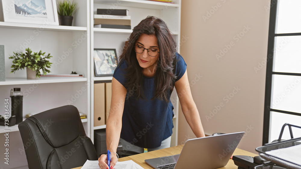 Professional hispanic woman in glasses writes in an office, surrounded by modern furnishings. Professional hispanic woman in glasses writes in an office, surrounded by modern furnishings.