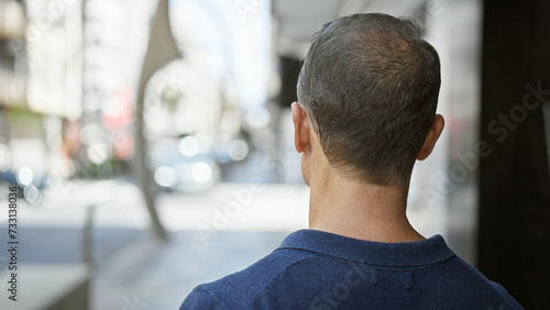 Handsome, mature hispanic man seen from behind, standing backward on an urban street, immersed in his thoughts while appreciating the city exterior