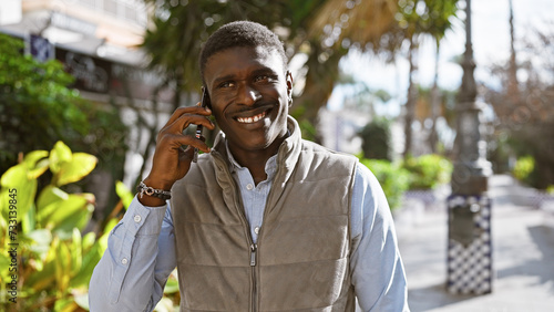 Smiling african man making a phone call outdoors in a sunny city park. © Krakenimages.com
