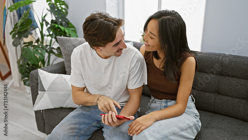 Interracial couple smiling together on a gray couch in a modern living room, sharing a loving moment indoors.