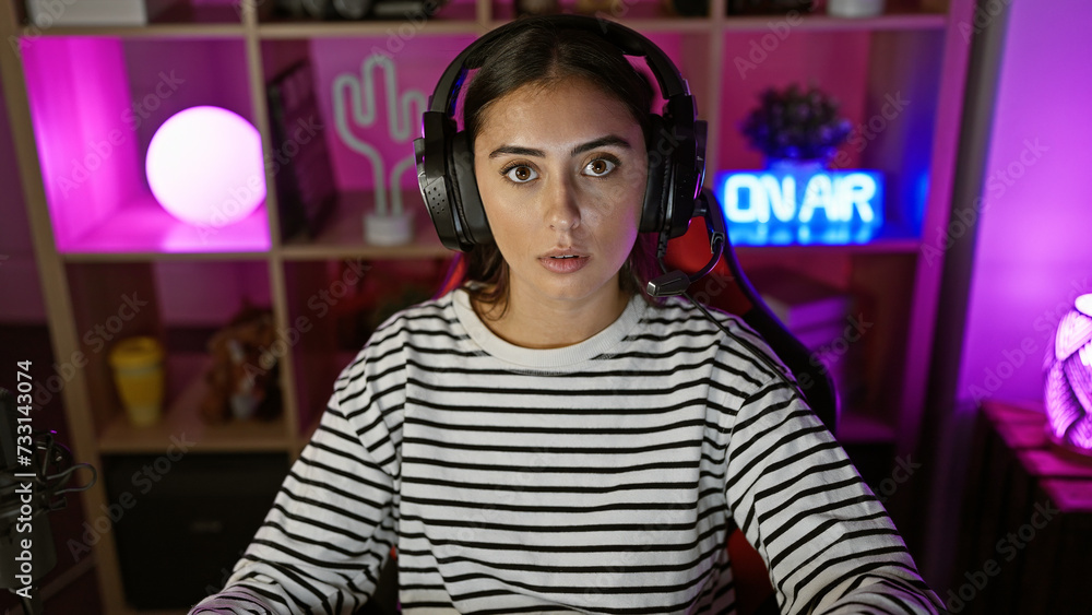 Fototapeta premium A young hispanic woman with headphones in a colorful gaming room looks surprised at night.