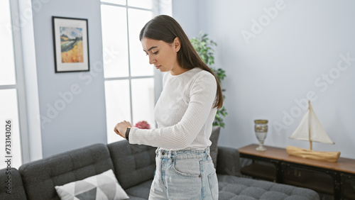 A young hispanic woman in casual attire checks the time indoors, suggesting punctuality in a modern home setting.