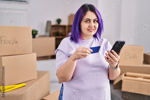 Young beautiful plus size woman using smartphone holding credit card at new home