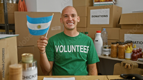 Young hispanic man volunteer smiling confident holding flag of argentina at charity center