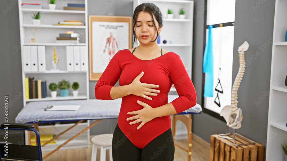 Hispanic woman practicing breathing exercises in a clinic interior with a visible spine model. Hispanic woman practicing breathing exercises in a clinic interior with a visible spine model.