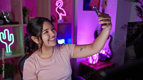 A smiling young hispanic woman takes a selfie in a colorful neon-lit gaming room at home during the evening. © Krakenimages.com