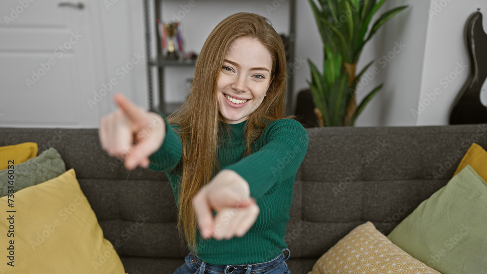 Caucasian blonde woman smiling and pointing at the camera in a cozy living room. Caucasian blonde woman smiling and pointing at the camera in a cozy living room.