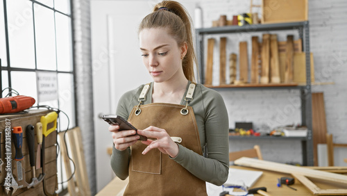 A focused young woman in a workshop, wearing an apron, using a smartphone amidst woodworking tools.