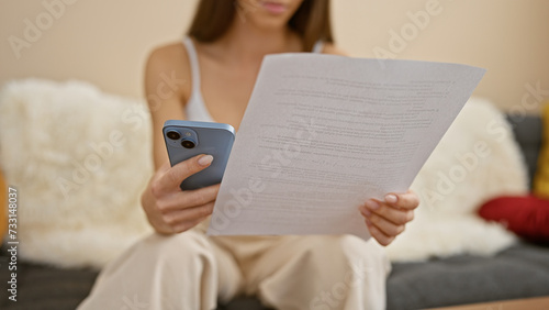 Smartphone in woman's hands, reading document with concentration while sitting on sofa at home, curious expression on her face; studying diligently, relaxed in her cozy living room.