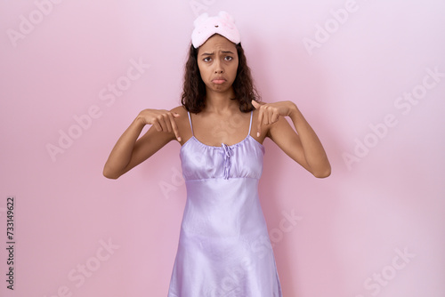 Young hispanic woman wearing sleep mask and nightgown pointing down looking sad and upset, indicating direction with fingers, unhappy and depressed.