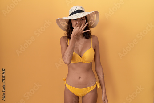 Young hispanic woman wearing bikini and summer hat smelling something stinky and disgusting  intolerable smell  holding breath with fingers on nose. bad smell