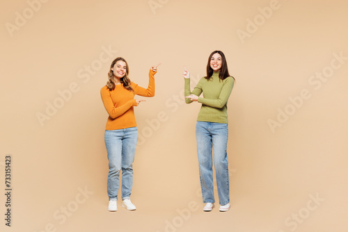 Full body young friends two women they wear orange green shirt casual clothes together point index finger on area between them isolated on plain pastel light beige background studio Lifestyle concept