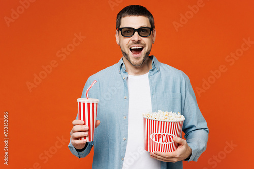 Young happy man he wears blue shirt white t-shirt casual clothes 3d glasses watch movie film hold bucket of popcorn cup of soda pop in cinema isolated on plain red orange background studio portrait.