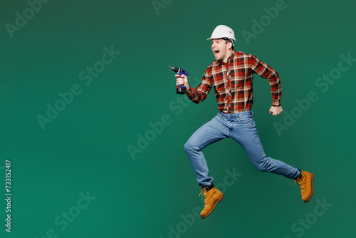 Full body young employee laborer handyman man wearing red shirt hardhat jump high with electric drill isolated on plain green background. Instruments for renovation apartment room Repair home concept photo