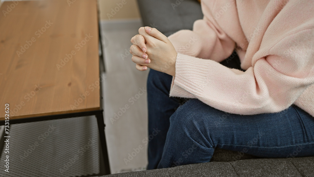 A contemplative young woman sits indoors, her hands clasped in a cozy, minimalist home setting.