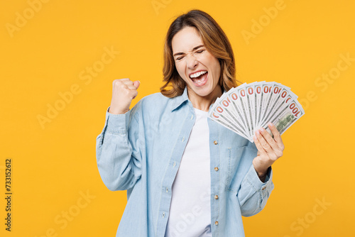 Young woman she wear blue shirt white t-shirt casual clothes hold in hand fan of cash money in dollar banknotes do winner gesture isolated on plain yellow background studio portrait Lifestyle concept photo