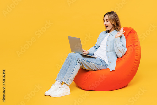Full body young IT woman wear blue shirt white t-shirt casual clothes sit in bag chair hold use work on laptop pc computer waving hand isolated on plain yellow background studio. Lifestyle concept.