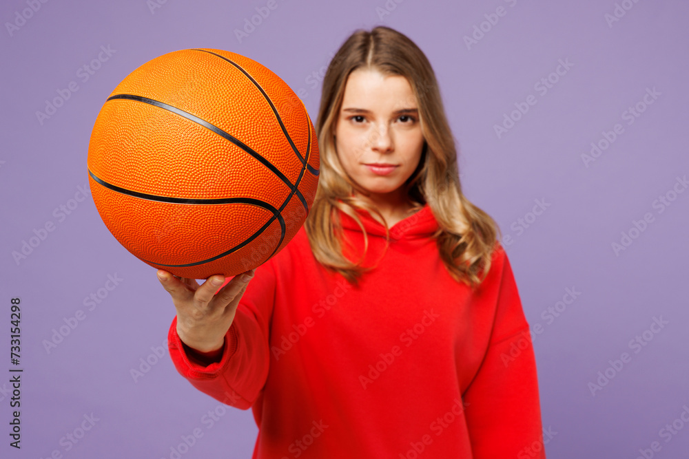 Young confident cool woman fan she wear red hoody cheer up basketball sport team hold in hand ball stretch hand to camera isolated on plain purple background. Sport leisure concept. Focus on object.