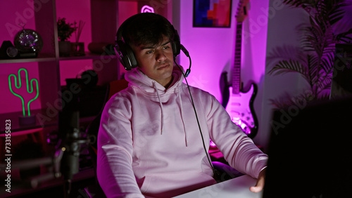 Focused young hispanic man curating a serious face, engrossed in streaming video games from a dark gaming room, sitting with headphones, plugged into the digital gaming world.