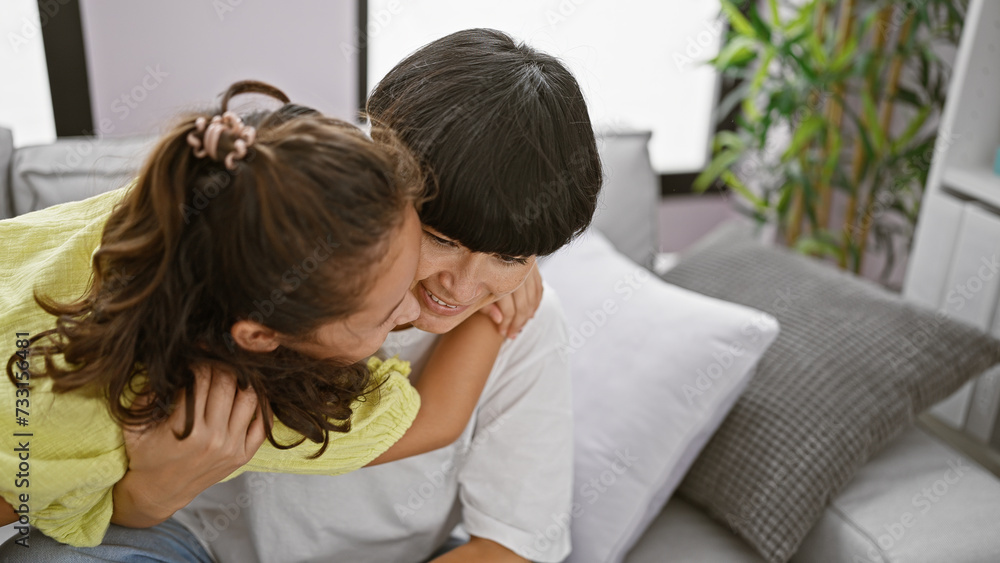 Confident mother and daughter sharing a joyful hug, smiling heartily while sitting on the living room sofa at home – a beautiful expression of family love and happiness Confident mother and daughter sharing a joyful hug, smiling heartily while sitting on the living room sofa at home – a beautiful expression of family love and happiness