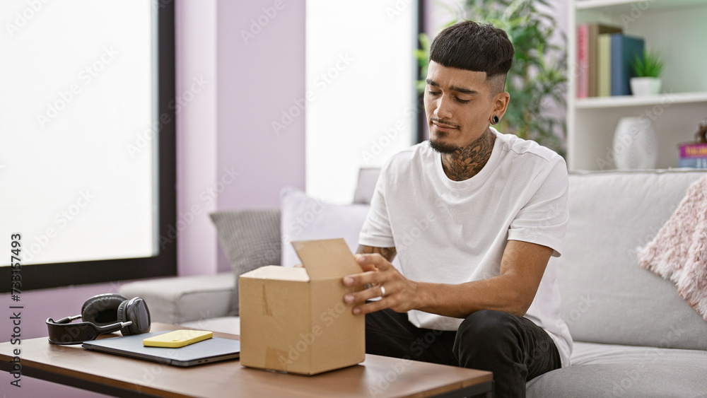 Serious young latin man immersed in unpacking a cardboard box at home, sitting relaxed on his sofa, tattooed face in deep concentration Serious young latin man immersed in unpacking a cardboard box at home, sitting relaxed on his sofa, tattooed face in deep concentration