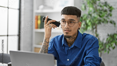 Focused latin worker, portrait of a handsome, tattooed business pro, serious and engrossed, working at his office, listening to an urgent voice message on his smartphone