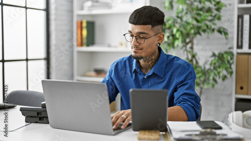 Concentrated young latin man at work, elegantly dressed, business professional operating a laptop and touchpad in his office environment. a serious, focused worker managing success, indoors. photo