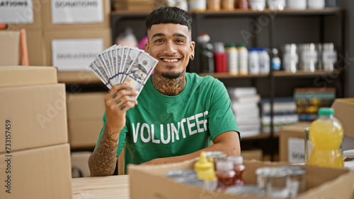 Handsome, tattooed young latin man, smiling confidently, volunteering at the charity center, holding dollars for donations in his hands