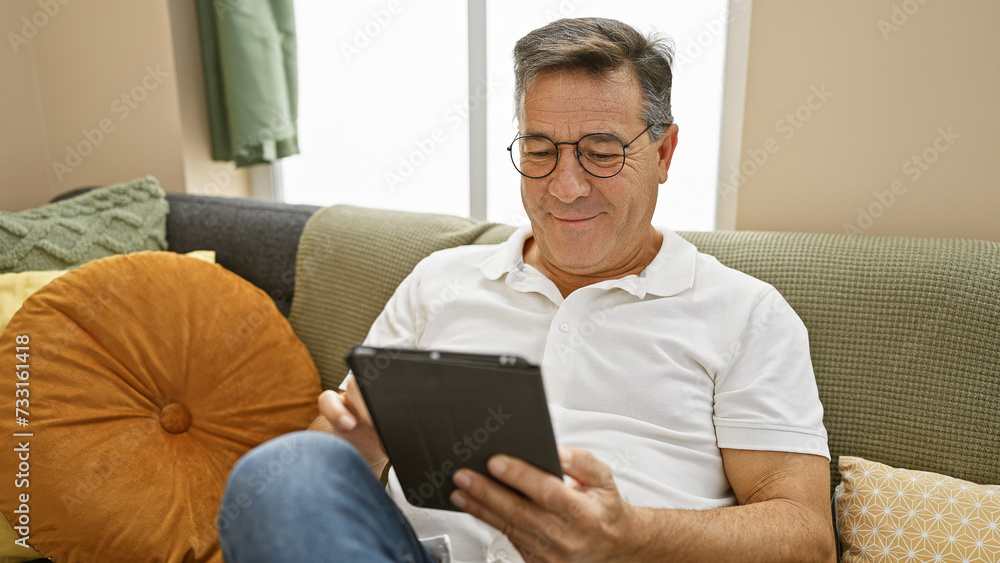 A middle-aged man relaxes at home, engaging with a tablet in his cozy living room. A middle-aged man relaxes at home, engaging with a tablet in his cozy living room.