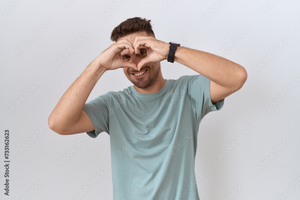 Hispanic man with beard standing over white background doing heart shape with hand and fingers smiling looking through sign Hispanic man with beard standing over white background doing heart shape with hand and fingers smiling looking through sign