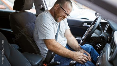 Stressed caucasian man in glasses, sitting in car on city street, suffering knee pain from driving photo