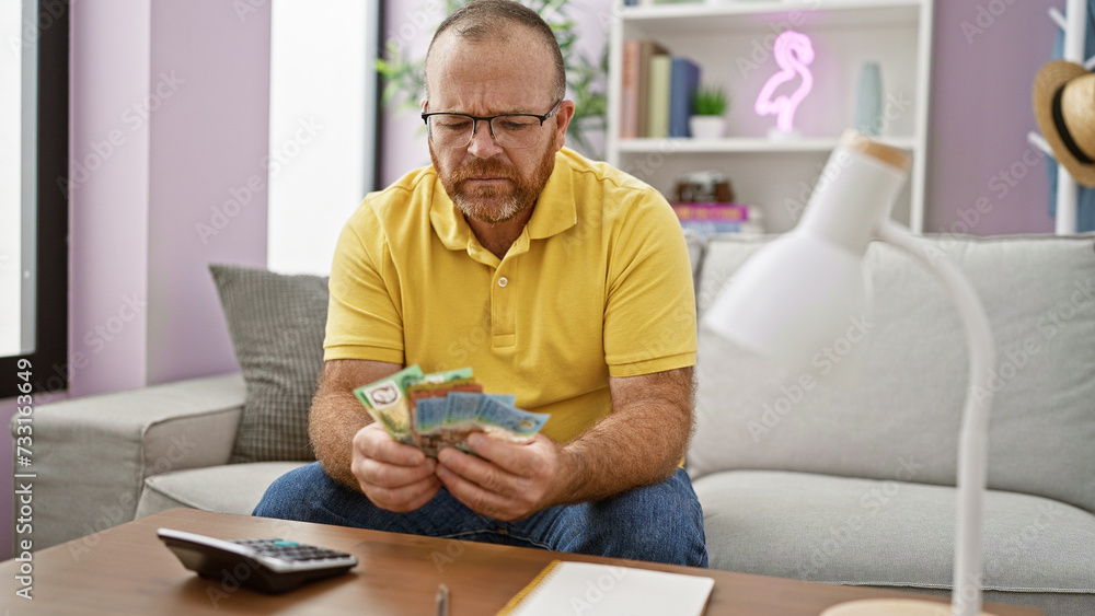 Handsome middle-aged caucasian man seriously sitting comfortably on home sofa, diligently counting australian dollars, marking careful finances in australia's economy - indoor adult living room scene.