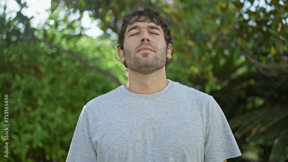 Hispanic young man with closed eyes, enjoying a breathing meditation in a sunny city park, a symbol of freedom and balance. blond beard, relaxed portrait, basking in the summer sunlight outdoors. Hispanic young man with closed eyes, enjoying a breathing meditation in a sunny city park, a symbol of freedom and balance. blond beard, relaxed portrait, basking in the summer sunlight outdoors.