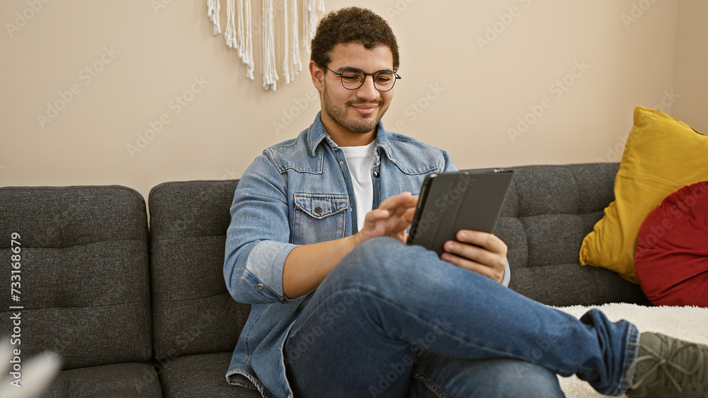 A young man with glasses and a beard enjoys using a tablet on a comfortable sofa indoors. A young man with glasses and a beard enjoys using a tablet on a comfortable sofa indoors.