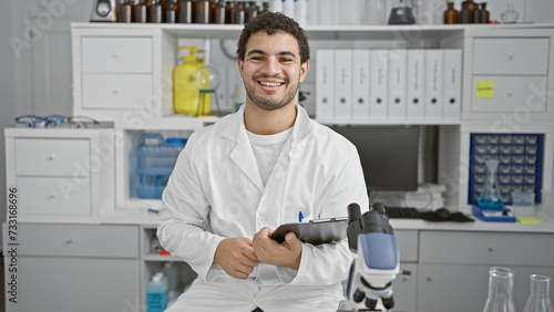 A smiling young man with a beard wearing a lab coat sits in a laboratory  writing on a clipboard.