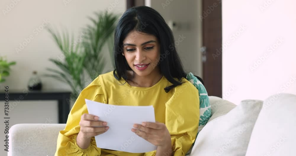 Joyful young Indian woman sit on couch, holds official paper document, read great news, received bank notice about loan approval, tax refund, get promotion, feel happy. Financial success, achievement Joyful young Indian woman sit on couch, holds official paper document, read great news, received bank notice about loan approval, tax refund, get promotion, feel happy. Financial success, achievement
