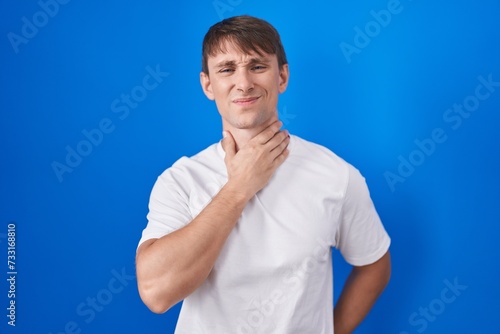 Caucasian blond man standing over blue background touching painful neck, sore throat for flu, clod and infection