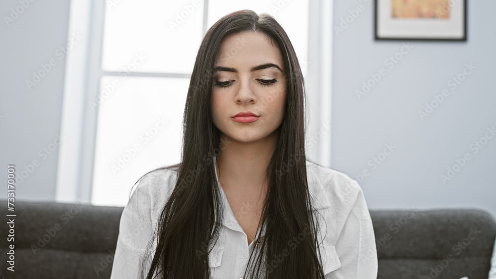 A serene young hispanic woman in a white blouse meditates indoors, providing a peaceful, attractive portrait scene.