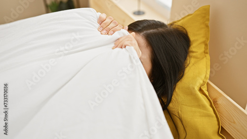 A young hispanic woman resting in an indoor bedroom with white bedding and a yellow pillow.
