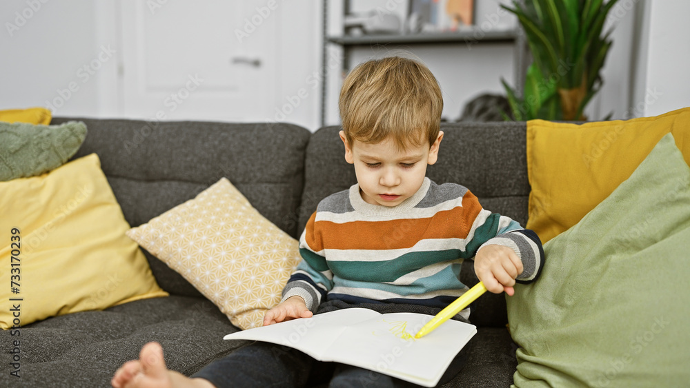 Caucasian toddler boy concentrating on drawing with a yellow crayon on white paper while seated comfortably on a gray couch indoors. Caucasian toddler boy concentrating on drawing with a yellow crayon on white paper while seated comfortably on a gray couch indoors.