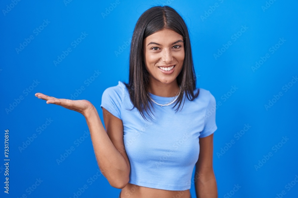 Brunette young woman standing over blue background smiling cheerful presenting and pointing with palm of hand looking at the camera. Brunette young woman standing over blue background smiling cheerful presenting and pointing with palm of hand looking at the camera.