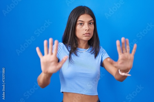 Brunette young woman standing over blue background moving away hands palms showing refusal and denial with afraid and disgusting expression. stop and forbidden.