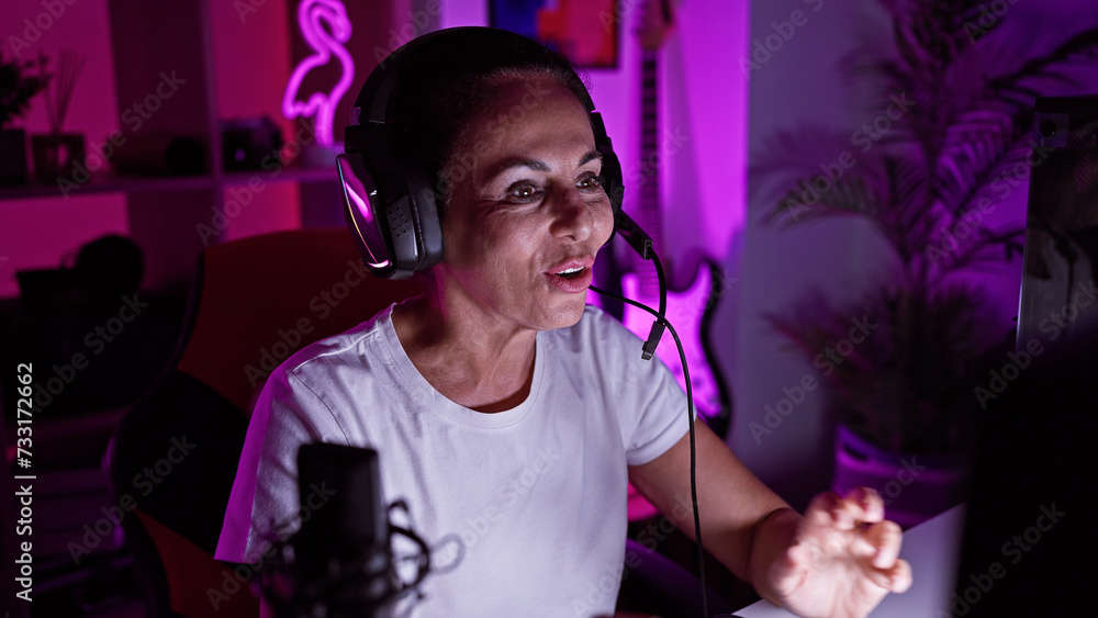 A middle-aged hispanic woman gaming at night in a vibrant home office set-up.