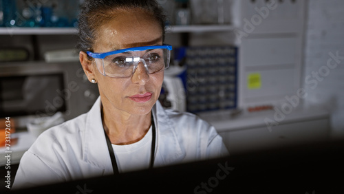 Hispanic woman researcher analyzes data intently in a laboratory setting, wearing safety goggles.